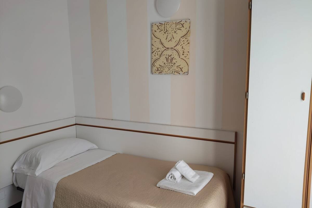 Een enkel bed met een beige dekbedovertrek en witte kussens, een opgevouwen handdoek op het bed, en een wandlamp.