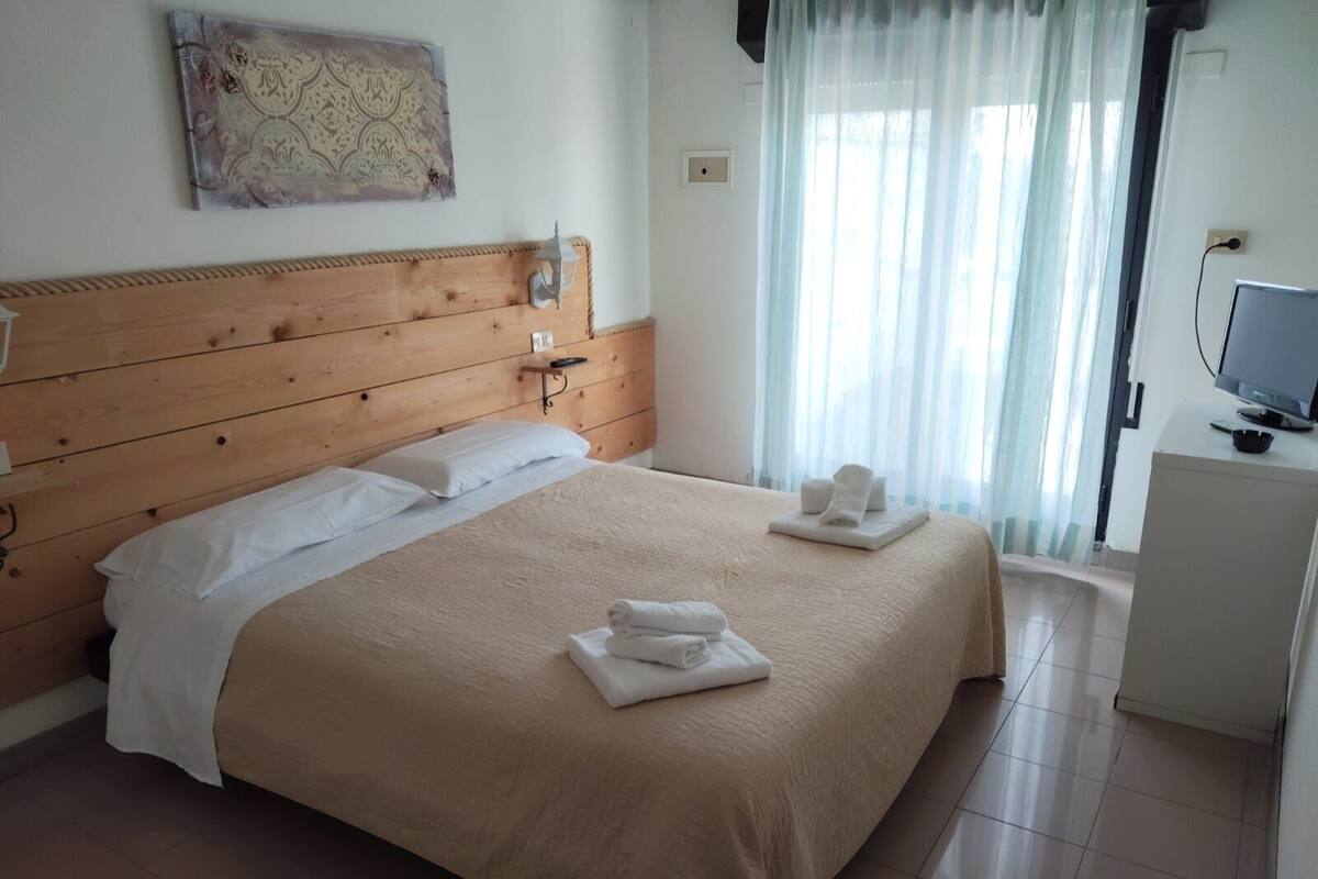 Een hotelkamer met een houten hoofdeinde, een bed met witte lakens, een flatscreen televisie op een witte kast en een raam met dunne gordijnen.