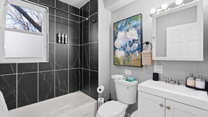 Un baño con bañera, inodoro y lavamanos. Hay una ventana con vistas a árboles y un cuadro de flores en la pared.