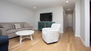 Un salon moderne avec un canapé, une table basse et un fauteuil à motifs. Un téléviseur à écran plat est fixé au mur, et on aperçoit un meuble de couleur turquoise à l’arrière-plan.