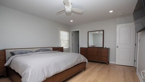Une chambre à coucher comprenant un lit, une commode en bois, un miroir, un ventilateur de plafond et une porte.