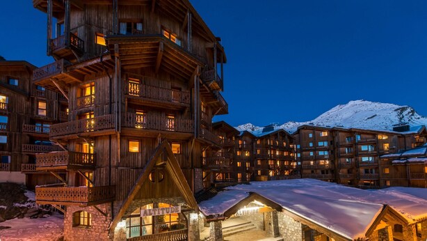 Un bâtiment en bois à plusieurs étages, doté de balcons et d’une base en pierre, avec une montagne enneigée en arrière-plan.