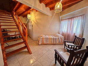 A room with a wooden staircase, a bed with striped bedding, two wooden chairs, and a window with curtains.