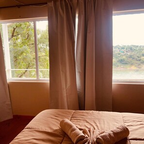 A room with a bed, a window with curtains, and a view of a river and greenery.