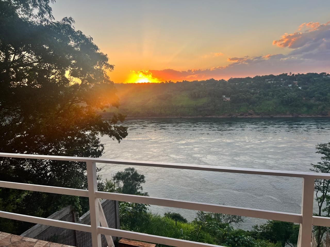 Um rio com o pôr do sol ao fundo, um corrimão em primeiro plano e vegetação nas margens.