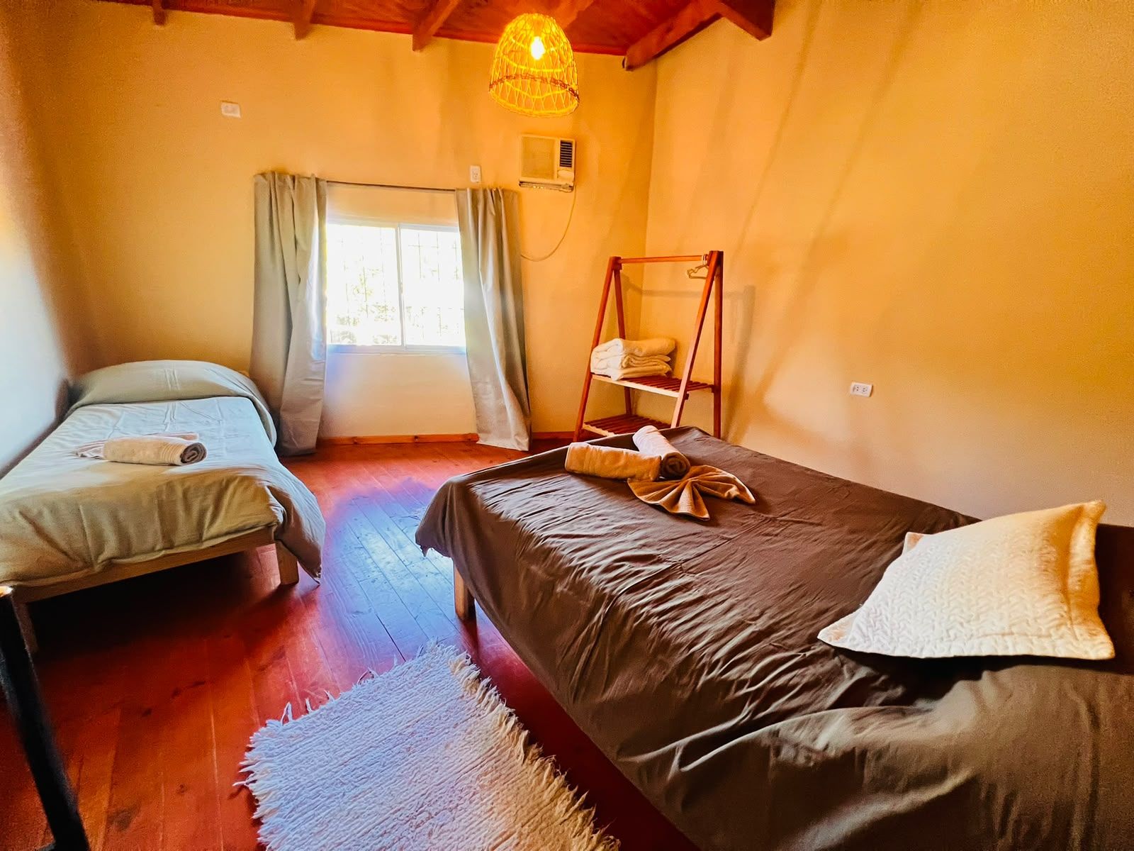 Una habitación con dos camas, piso de madera, ventana con cortinas, lámpara colgante y un estante con toallas.