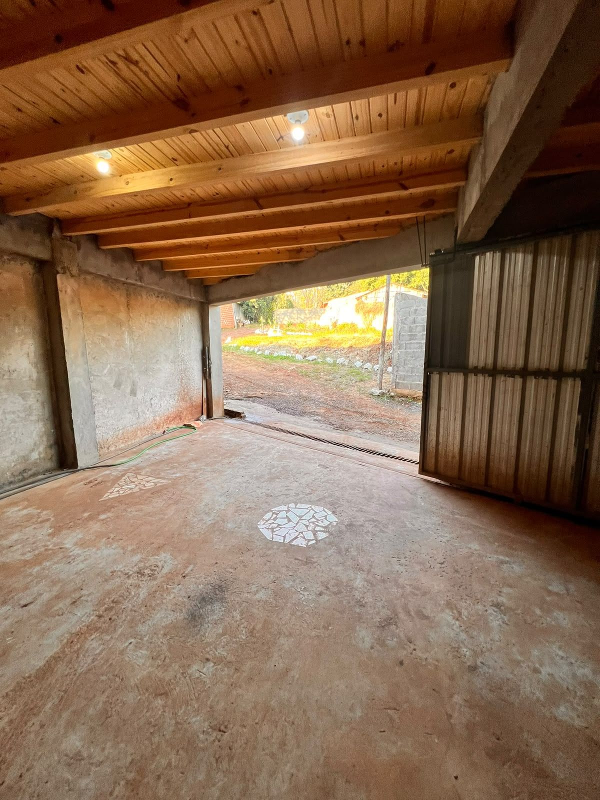 Un ambiente interno con pavimento in cemento, travi a vista in legno e vista su uno spazio esterno con muro in mattoni e vegetazione.