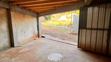 Un ambiente interno con pavimento in cemento, travi a vista in legno e vista su uno spazio esterno con muro in mattoni e vegetazione.