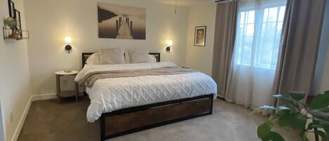 A bedroom with a bed, a ceiling fan, a nightstand, a wall-mounted lamp, a window with curtains, and a potted plant.