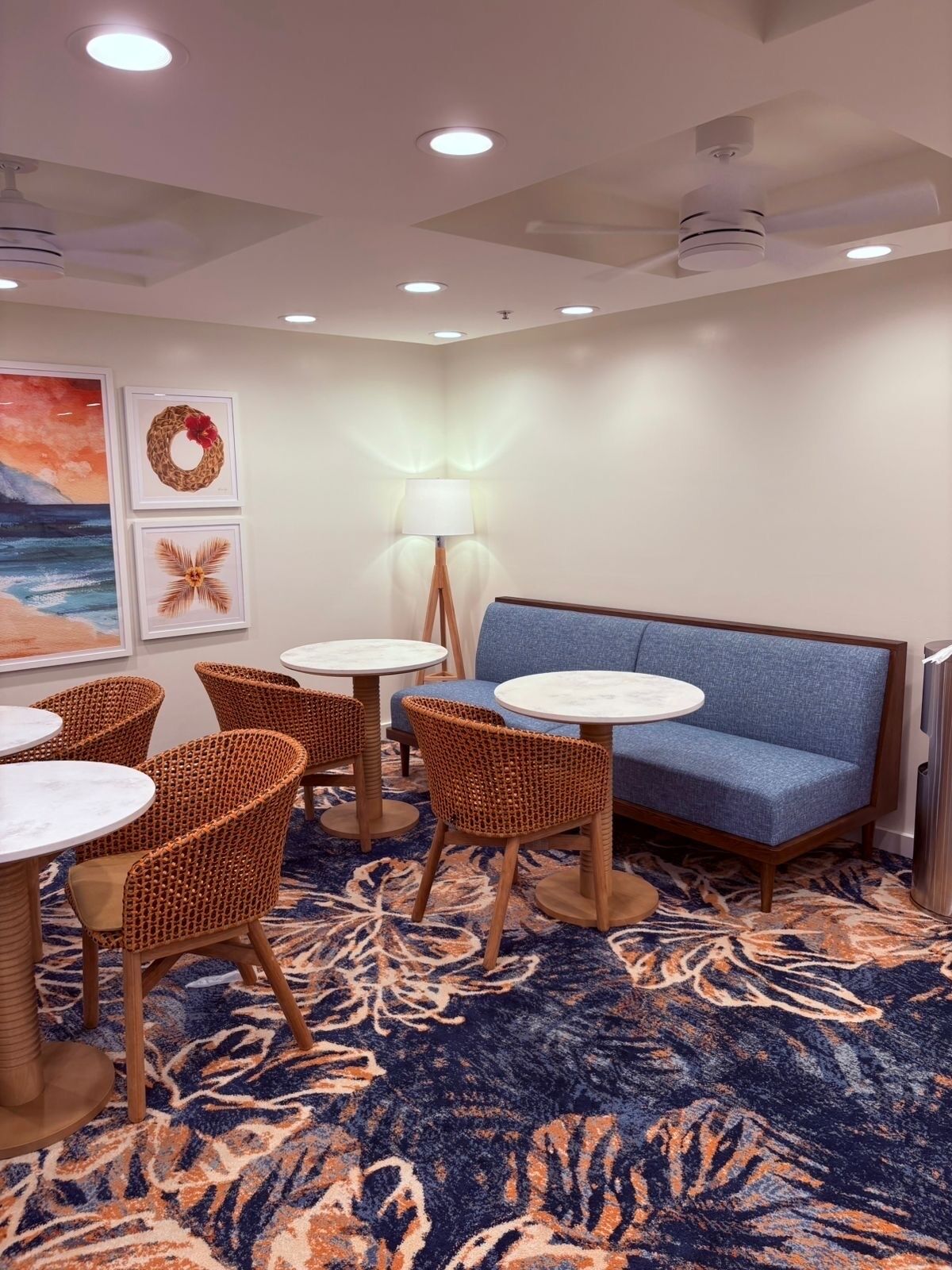 A room with a blue sofa, wicker chairs, round tables, a patterned carpet, and wall art.
