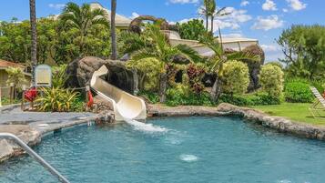 A pool with a water slide, surrounded by tropical plants and palm trees.