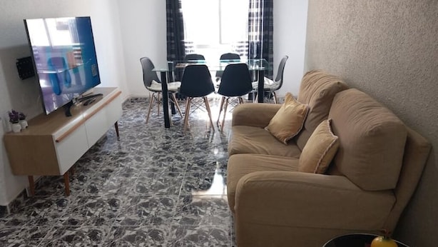 Una sala de estar con un sofá color beige, un televisor de pantalla plana, una mesa de comedor de vidrio con sillas y un piso de mármol.