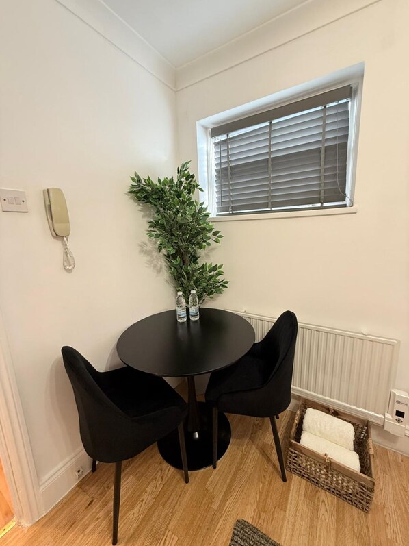 Quarto de canto com uma mesa redonda, duas cadeiras pretas, um vaso de planta, uma janela com persianas, um telefone, um radiador e uma cesta de vime com toalhas.