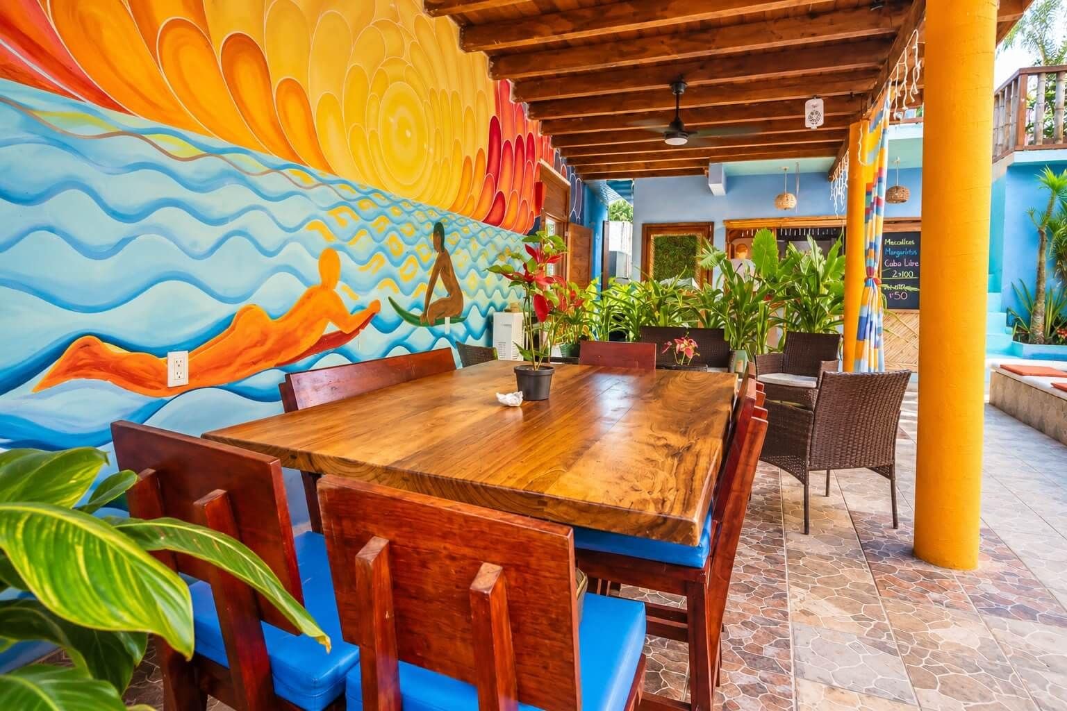 A wooden dining table with blue cushioned chairs, surrounded by potted plants and a vibrant mural.