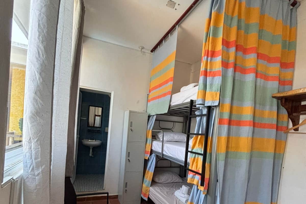 Een studentenkamer met stapelbedden, een wastafel, een klein tafeltje en een raam met gordijnen.