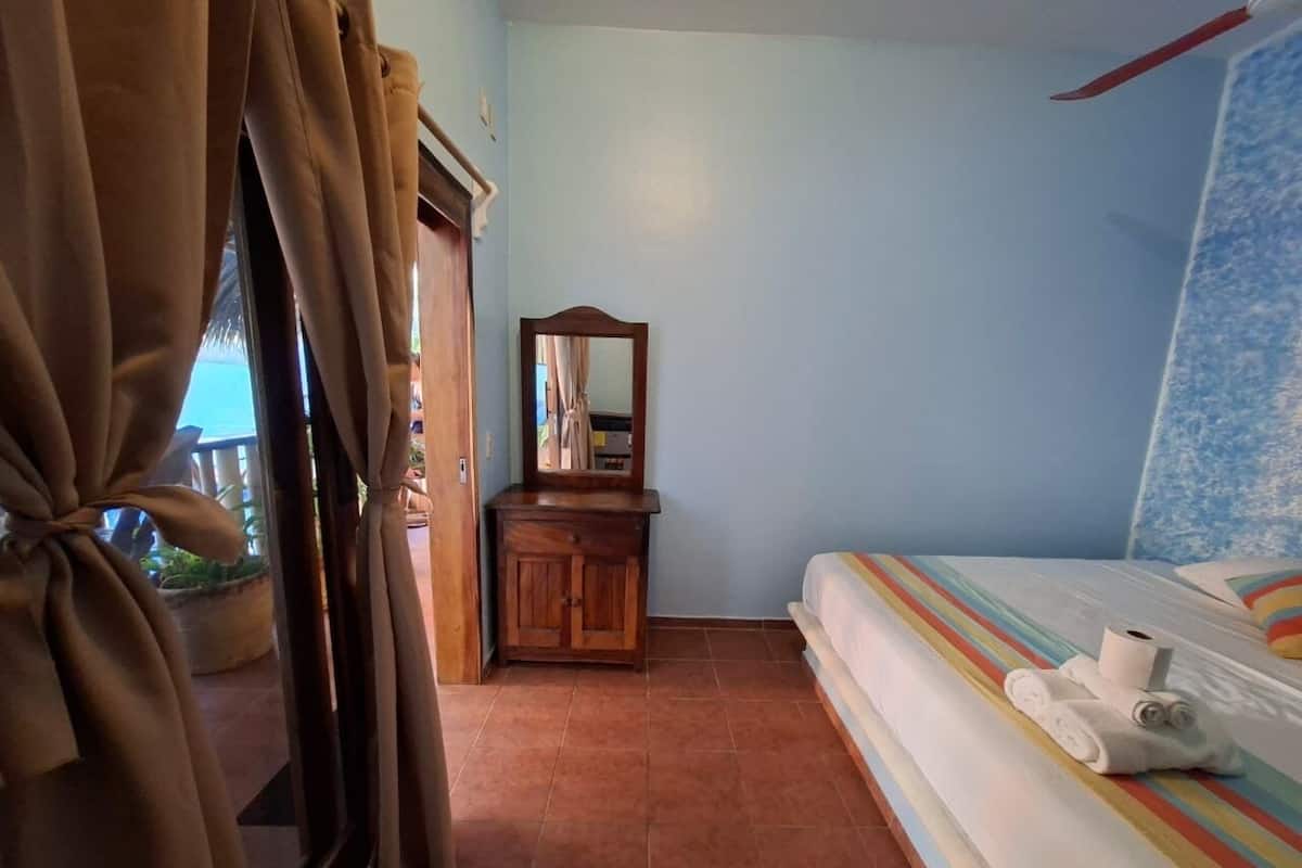Een slaapkamer met een bed, een houten dressoir, een spiegel en uitzicht door een raam met gordijnen.