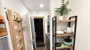 Un couloir étroit avec une armoire en bois, une chaise noire et une étagère où sont posés de la vaisselle et un pot de fleurs.