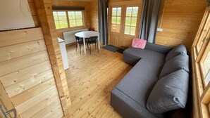 Interior de una cabaña de madera con un sofá gris, una mesa de comedor con sillas y ventanas grandes.