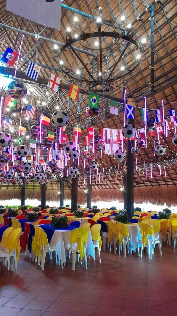 Un gran salón con mesas cubiertas con manteles amarillos y blancos, rodeado de sillas. Banderas y balones de fútbol cuelgan del techo.