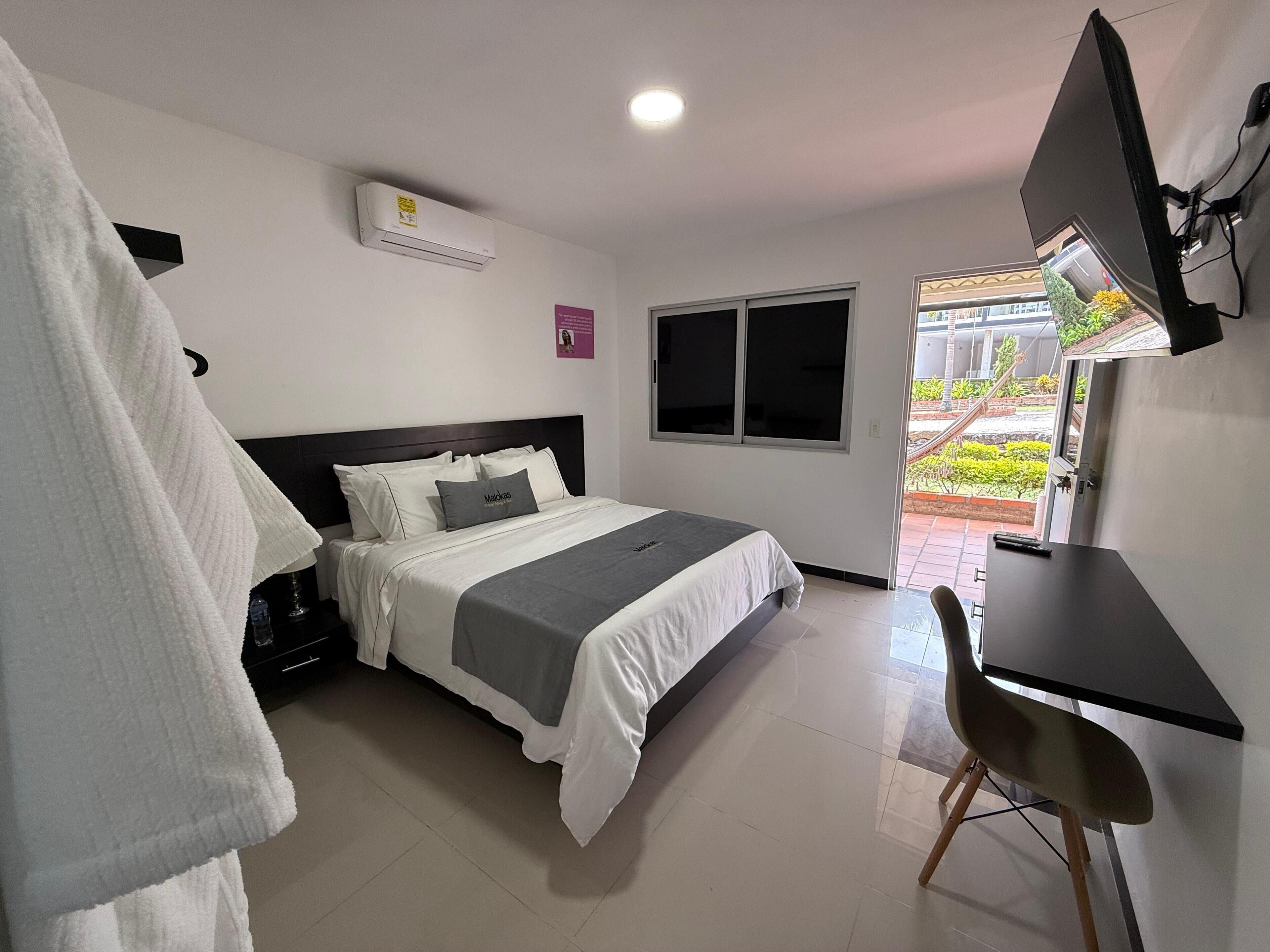 A bedroom with a bed, a desk, a chair, a TV, and a window.