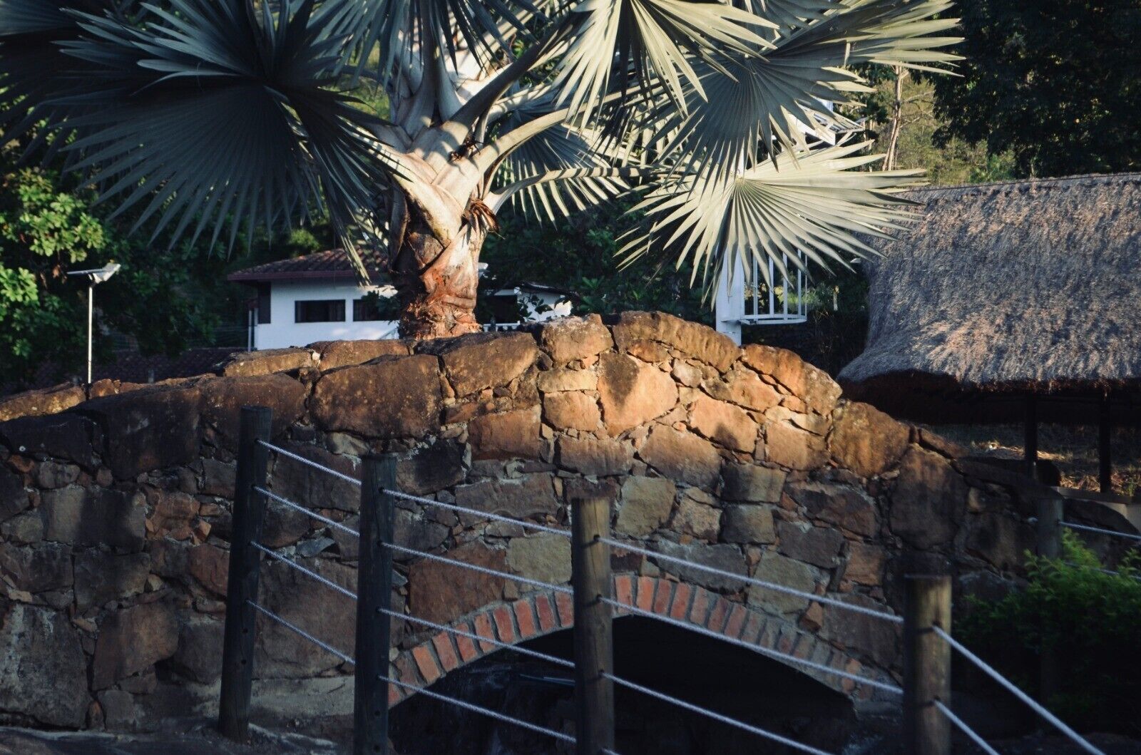 Un pont en pierre surmonté d’une structure au toit de chaume, entouré d’une végétation luxuriante et d’un palmier.