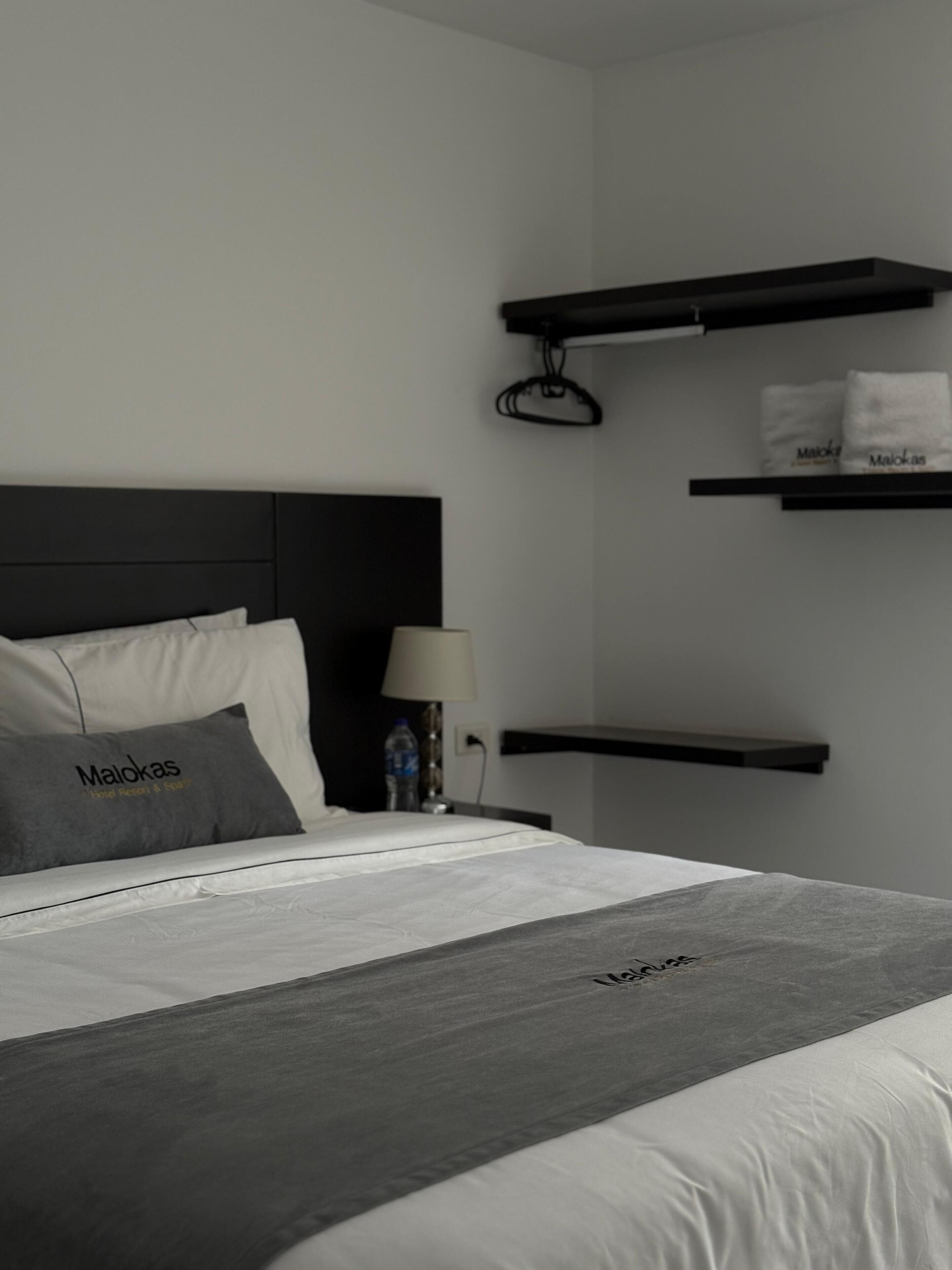 A hotel room with a bed, pillows, a bedside table with a lamp, and two wall-mounted shelves with towels.