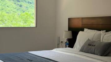 A hotel room with a large window, a bed with white linens, a dark headboard, and a nightstand with a lamp and a bottle.