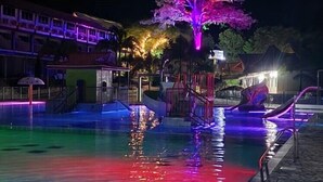 A nighttime swimming pool with colorful lighting, a tree illuminated in purple, and a building in the background. - MALOKAS RESORT (Socorro)