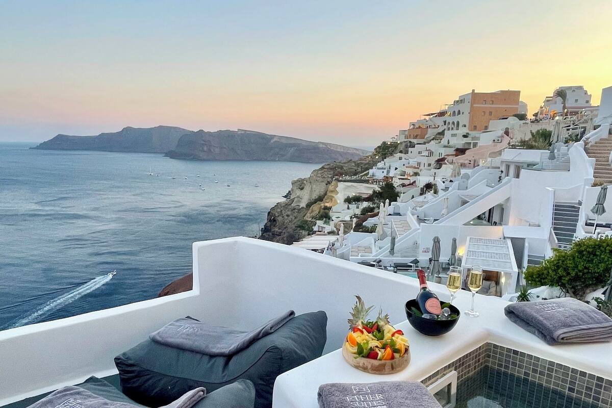 A rooftop terrace with white walls, grey cushions, a small pool, and a sunset view of a coastal town.