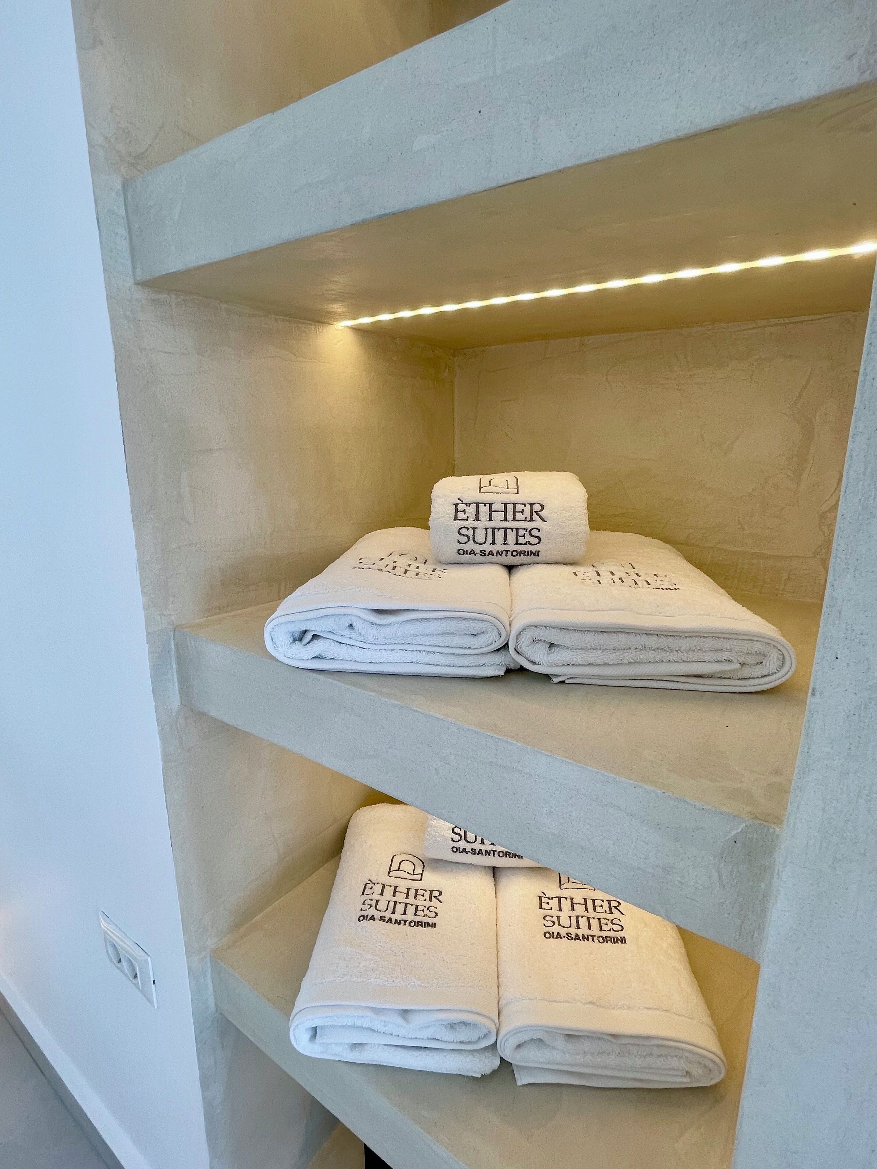 Ein Hotelzimmer mit einer Ablage, auf der ordentlich gefaltete Handtücher liegen, beschriftet mit 'ÉTHER SUITES OIA-SANTORINI'.