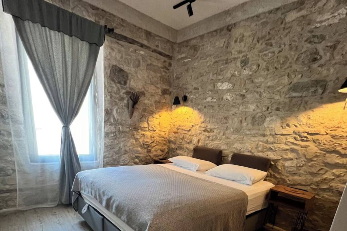 A bedroom with a stone wall, a bed with a gray blanket, a small table, and a window with curtains.