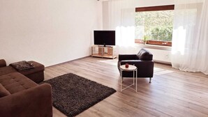 Un salon moderne doté d’un téléviseur à écran plat, d’un canapé, d’une table basse et d’une grande fenêtre agrémentée de rideaux.