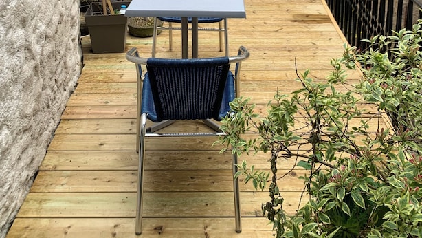 Una terraza de madera con una mesa y sillas, rodeada de vegetación y muros de piedra.