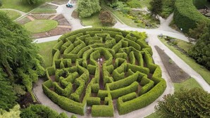 Un grand labyrinthe circulaire formé de haies, avec un chemin central menant à un petit pont qui enjambe un élément décoratif aquatique.