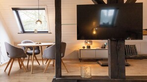 A modern living room with a television, wooden beams, a dining table with chairs, and a window with a view of trees. - Lütt Heidjer Hus- Stylish oasis of peace. Guests with pets welcome (Buchholz in der Nordheide)