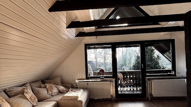 A spacious attic room with a wooden slatted ceiling, a large window with a view of trees, a sofa, a coffee table, and a radiator. - Lütt Heidjer Hus- Stylish oasis of peace. Guests with pets welcome (Buchholz in der Nordheide)