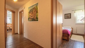 Ein Flur mit Holzboden, einem Hausbild an der Wand, einem Bett mit rosa Decke und einem Fenster mit Vorhang.