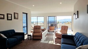 A spacious living room with a blue sofa, two recliners, and a coffee table. Large windows offer a view of the city and mountains. - A large and sunny family home close to all the amenities and perfect for exploring Nelson and Tasman (Richmond)