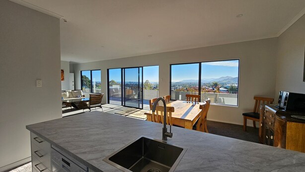 A modern kitchen with a large island, a dining area with a view, and a living space with a sofa and table. - A large and sunny family home close to all the amenities and perfect for exploring Nelson and Tasman (Richmond)