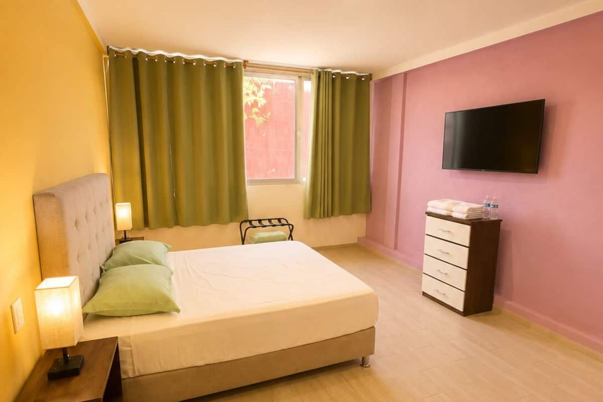 A bedroom with a bed, a nightstand, a dresser, a TV, and a window with curtains.