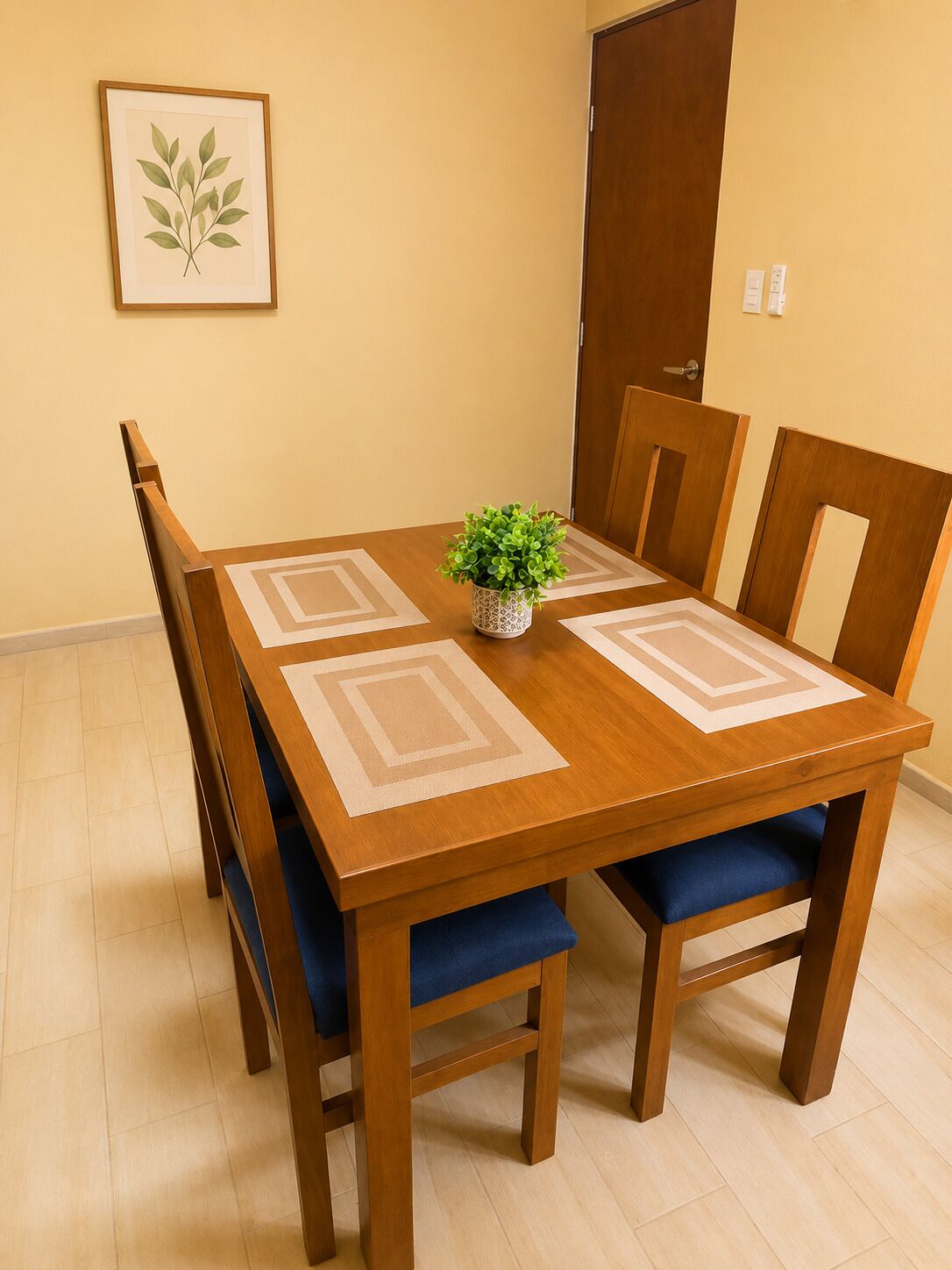 Een houten eettafel met blauwe stoelen met kussens, een kleine potplant en een ingelijst botanisch kunstwerk aan de muur.