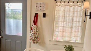 A room with a gray door, a white wall clock, a window with a striped curtain, two white chairs, and a small table with a plant. - The Garden Alley View (Arcata)