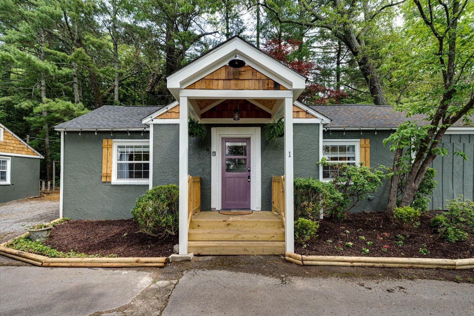 A small, single-story house with a wooden porch and a purple door, surrounded by a garden and trees.