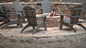 Un espace extérieur accueillant, aménagé avec des fauteuils Adirondack et un foyer en pierre, entouré d’un muret en pierre et d’un petit bâtiment en bois doté d’une véranda.