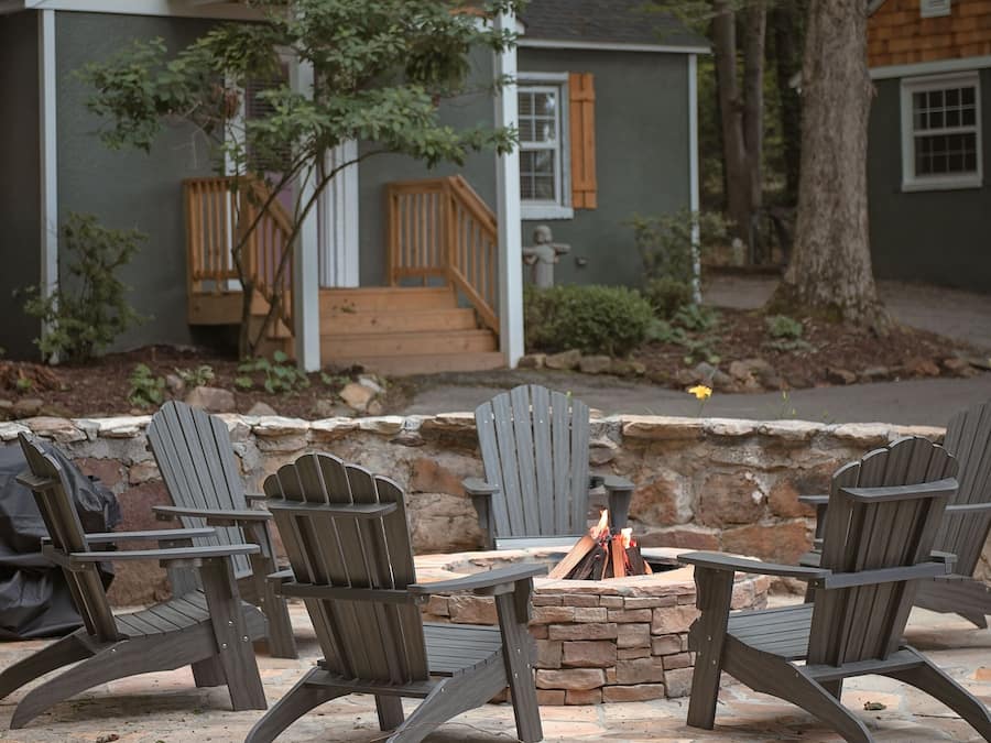Un espace extérieur accueillant, aménagé avec des fauteuils Adirondack et un foyer en pierre, entouré d’un muret en pierre et d’un petit bâtiment en bois doté d’une véranda.