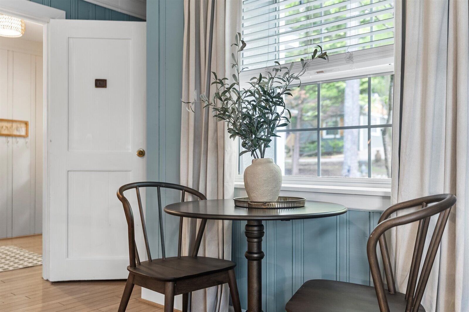 A room with a round table, two chairs, a vase with a plant, and a window with blinds.