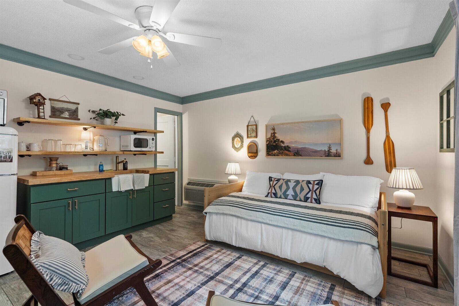 A bedroom with a bed, bedside tables, a chair, a desk, a kitchenette, and a ceiling fan.