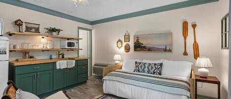 A bedroom with a bed, bedside tables, a chair, a desk, a kitchenette, and a ceiling fan.