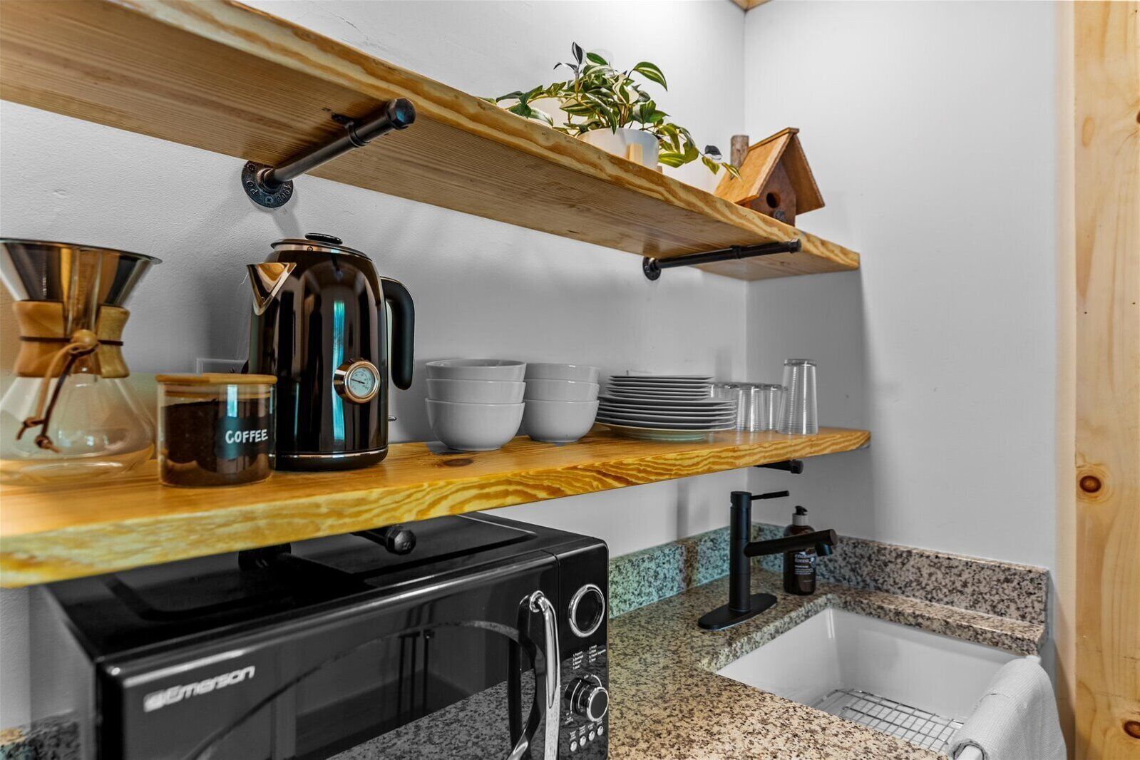 A compact kitchen with a coffee maker, a kettle, and a microwave on the counter. There is a wooden shelf above the counter holding a plant and a birdhouse.
