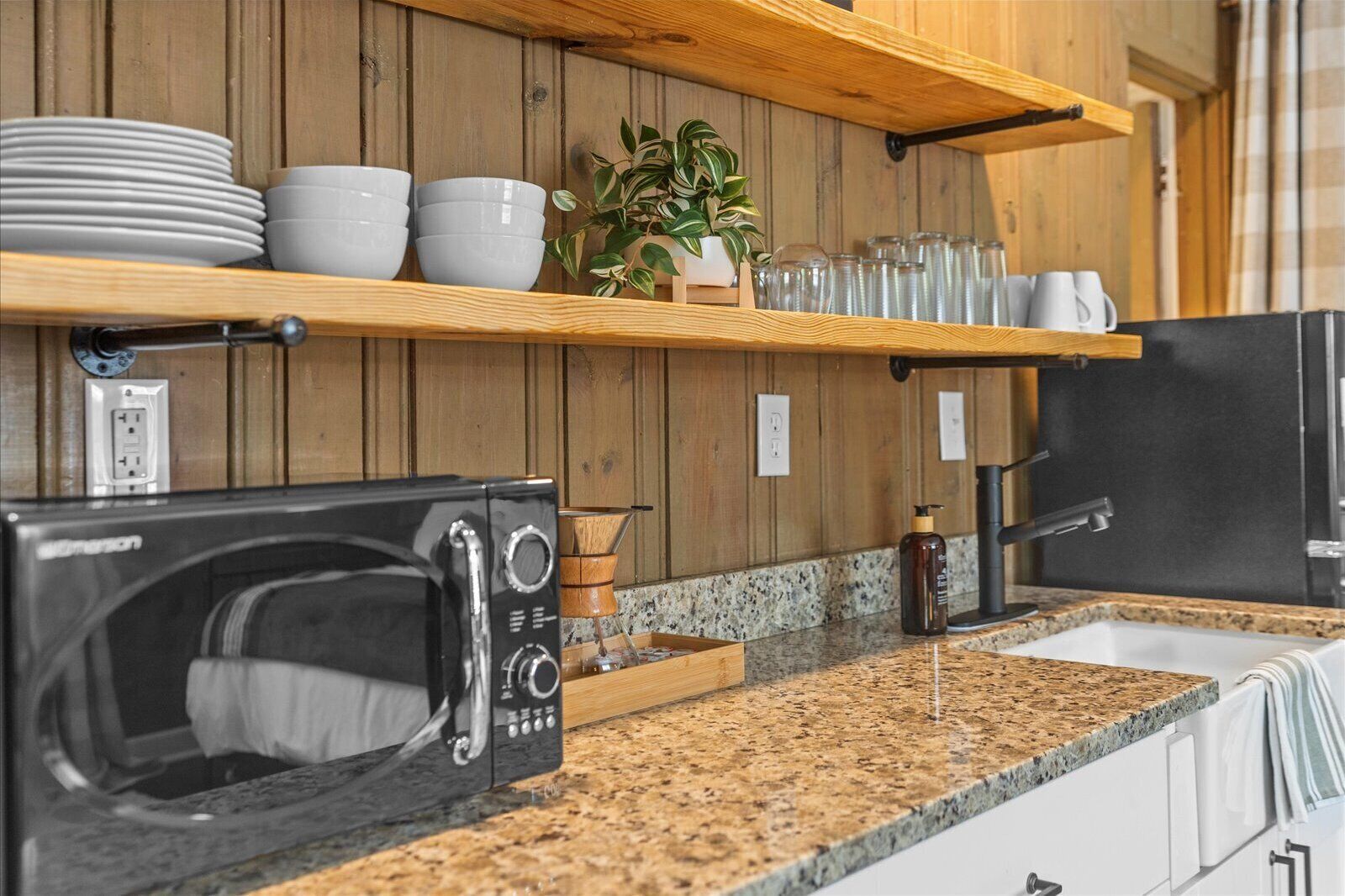 Una cocina con encimeras de granito, un microondas negro, un fregadero y estantes de madera con platos y vasos.